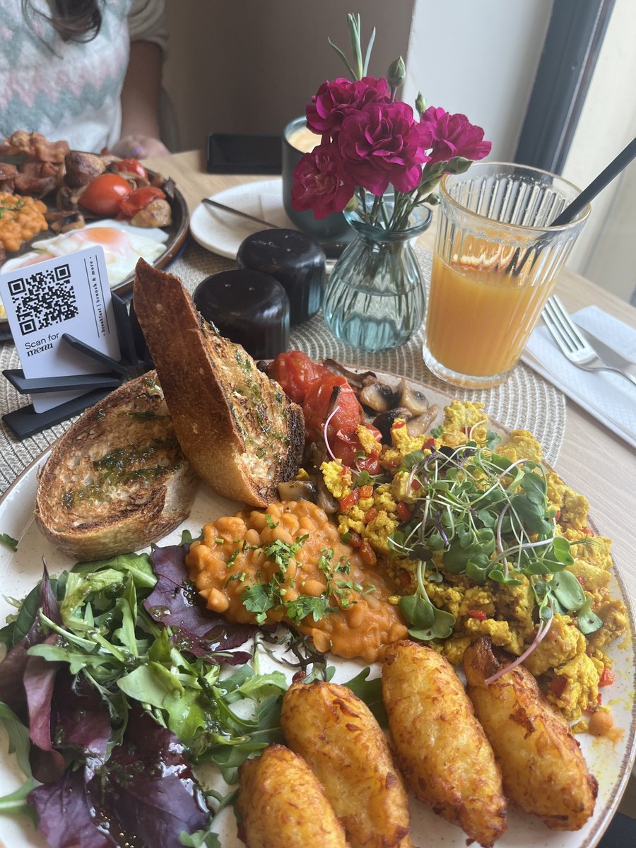 Een brunchbord met tofu scramble, toast, bonen, groene salade en gebakken aardappel, met sap op de achtergrond.