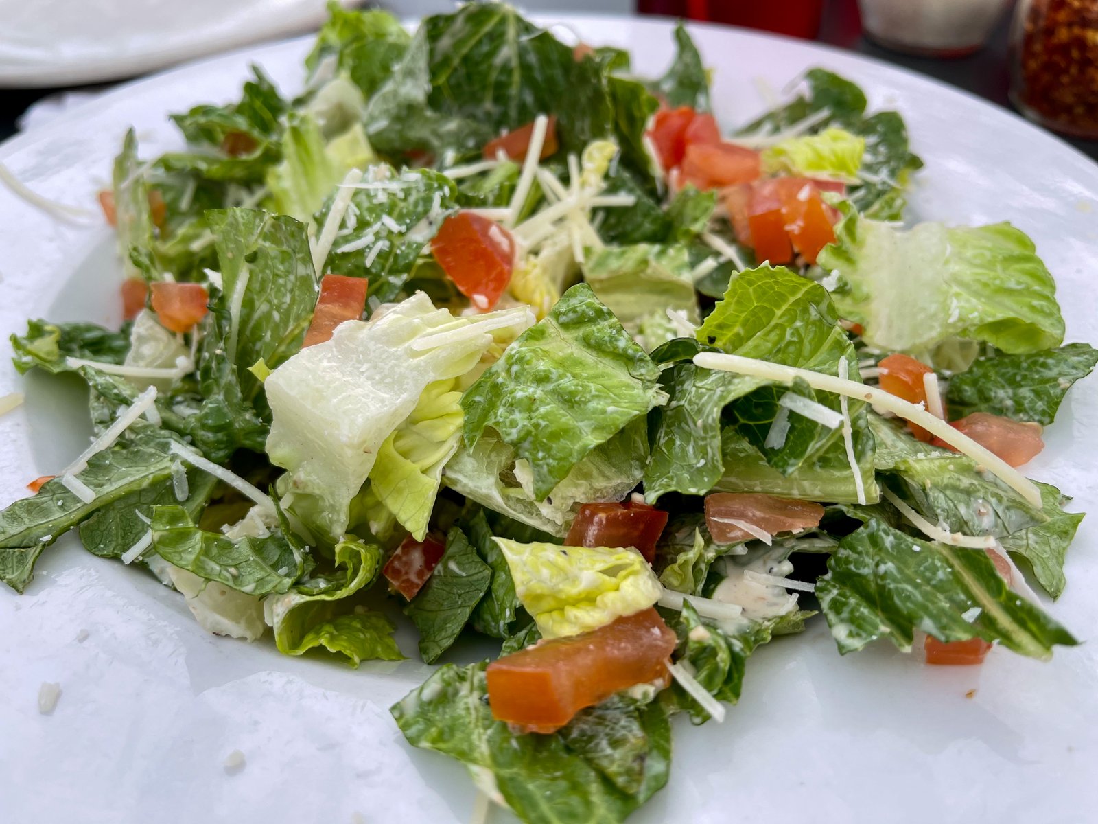 Een salade met groene bladeren, tomaat en geraspte kaas op een wit bord.